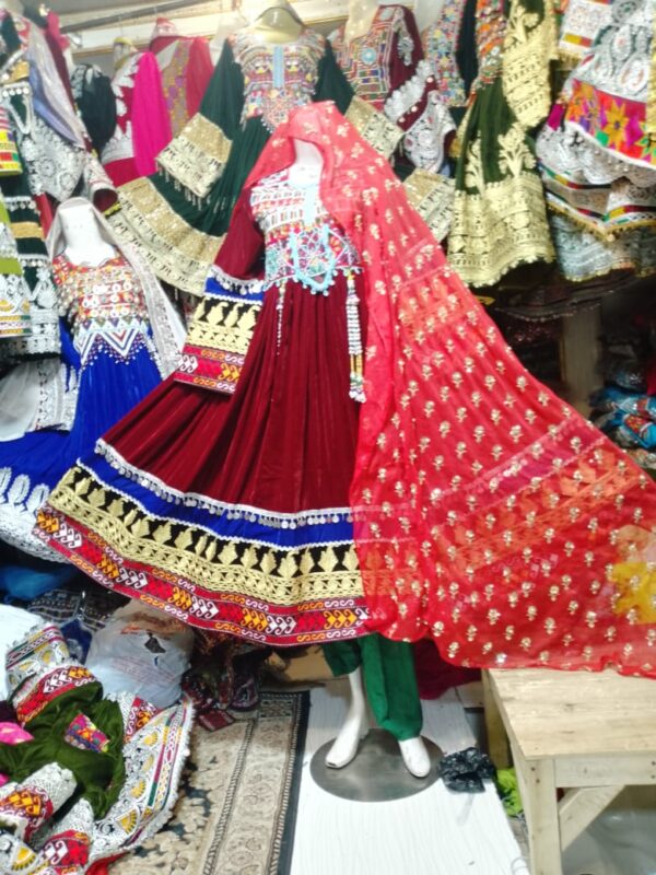 Maroon velvet Afghani frock with multicolor embroidery and golden threadwork, displayed on a mannequin with a sheer red dupatta and surrounded by traditional Afghan garments in a cultural boutique.