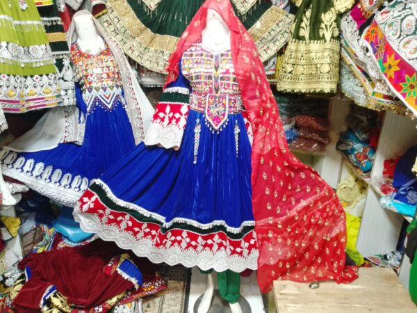 "Blue Velvet Afghani Frock adorned with vibrant traditional embroidery in red, white, and green, featuring a full skirt with white lace hem and intricate border detailing. A red dupatta with golden accents complements the rich design. Set against a backdrop of cultural garments, the frock showcases Afghan heritage and handcrafted artistry."