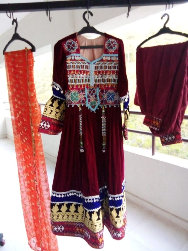 Velvet Afghani Dress for Cultural Celebrations displayed on hangers, featuring a richly embroidered maroon outfit with intricate patterns, an orange floral scarf, and matching velvet pants—showcasing traditional Afghan craftsmanship and festive elegance.
