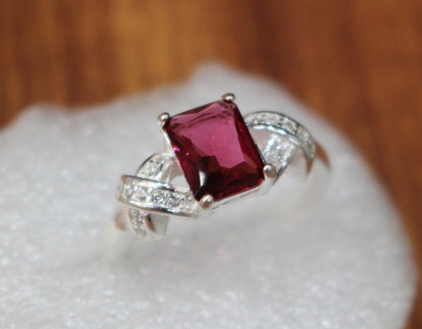 A close-up of a ruby ring featuring a vibrant, square-cut ruby set in rhodium-plated silver. The band showcases elegant detailing, and the ring rests on a soft white cushion with a warm wooden background.