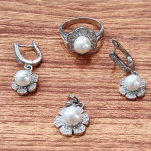 A handcrafted freshwater pearl jewelry set featuring a floral-themed ring, elegant drop earrings, and matching pendants. Each piece is adorned with a luminous freshwater pearl nestled in a rhodium silver flower design, accented with delicate shimmering stones—displayed on a warm wooden surface that highlights its artisanal charm.