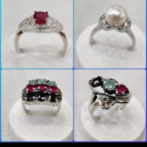 “Four distinct gemstones engagement rings displayed on white circular stands: one with a central ruby and clear accents, one featuring a large pearl surrounded by shimmering stones, one arranged in a rectangular cluster of red, green, and black gemstones, and one showcasing paired red and green stones with intricate silver detailing.”