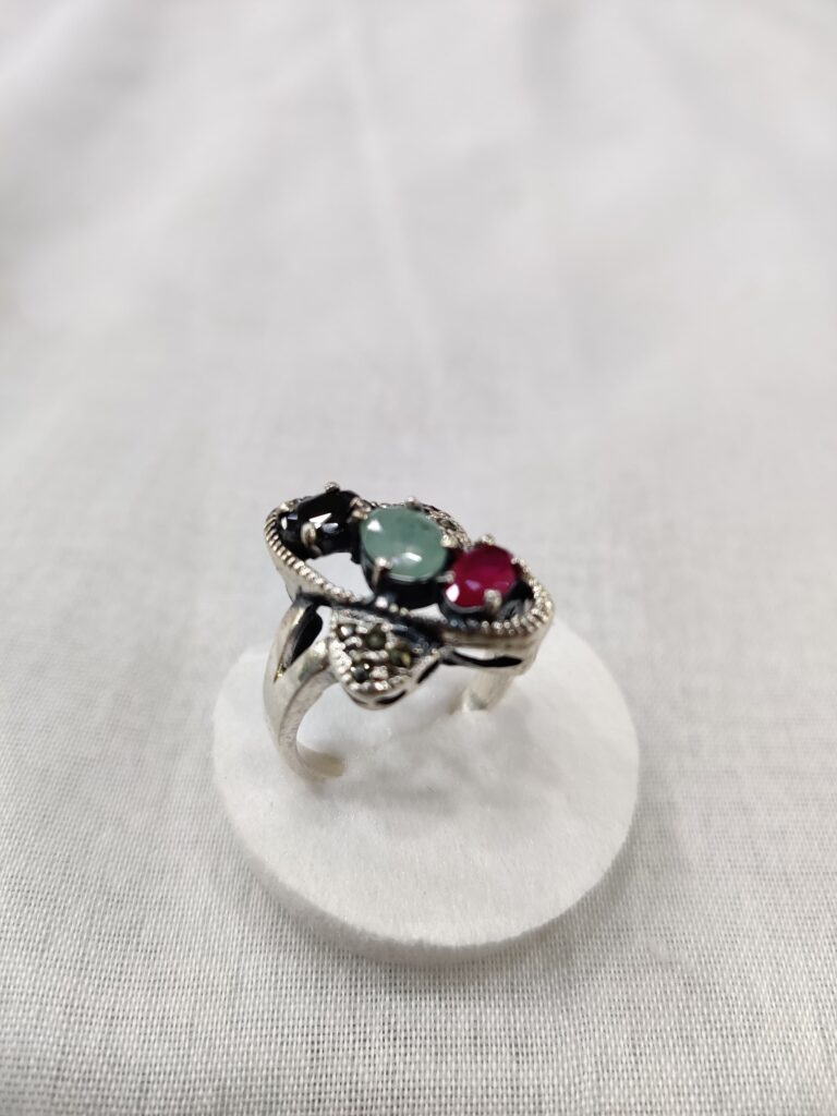 Silver ring with green and red gemstones, elegantly set in an intricate design, displayed atop a white circular stand against a soft gray background.
