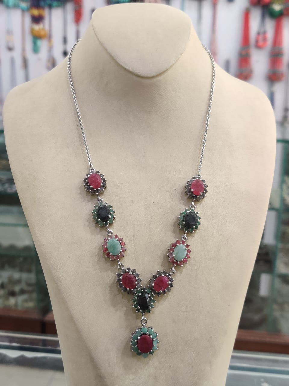 "Silver necklace with red, green, and blue gemstone clusters arranged in a V-shape, displayed on a beige mannequin bust in a jewelry store."