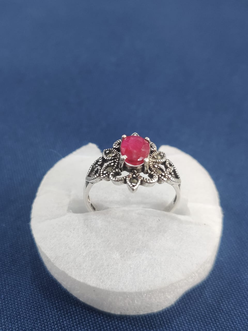 "Ornate silver ring with a central red gemstone, displayed on a white cushion against a blue fabric background, featuring floral detailing and vintage Afghan craftsmanship."